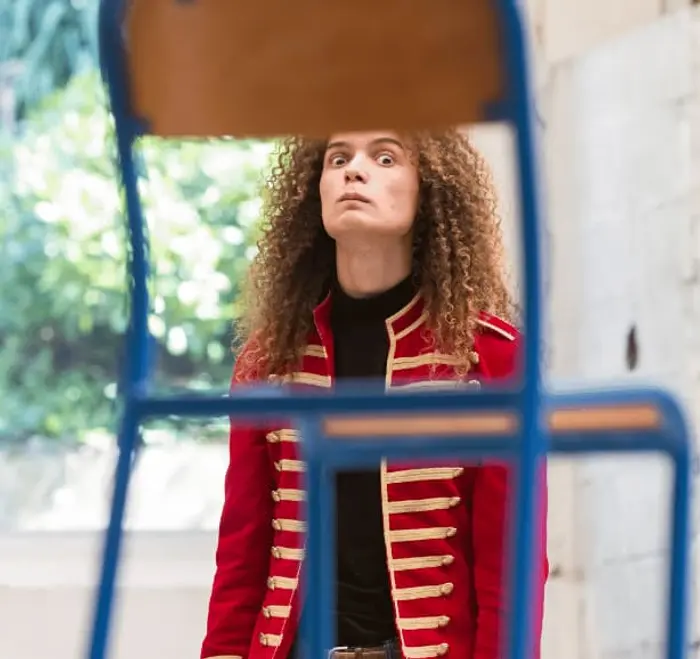 Un homme nous fixe de ses grands yeux au travers une chaise d'école entre le dossier et l'assise qui est au premier plan. 
