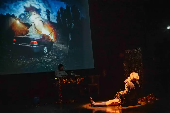 Une femme est assise sur la scène. Elle a le regard levé vers un écran sur lequel passe une image de voiture en feu qui rentre dans une maison. Sous cette projection se trouve un DJ, assis à sa table.