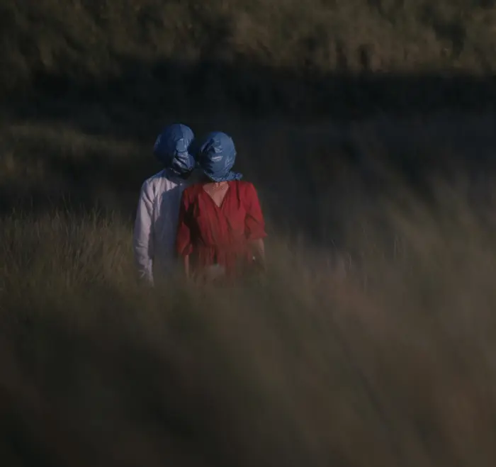 Dans un champ avec des herbes hautes, 2 personnes se tiennent debout. Elles sont très proches, reliées entre elles par un sac plastique bleu qui recouvre leurs têtes. La femme, vêtue d'une robe rouge, se trouve juste devant l'homme,  qui porte une chemise blanche.
