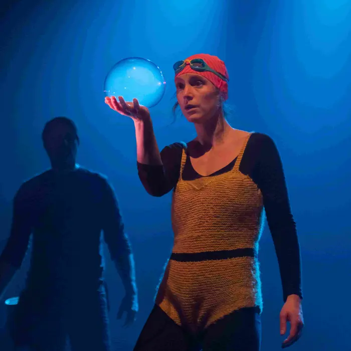 Camille Demoures est sur scène. Elle porte un bonnet de bain et des lunettes de piscine, ainsi qu'un ensemble en laine jaune. Elle porte avec sa main droite une sorte de grosse boule d'eau. Un homme se tient derrière elle.