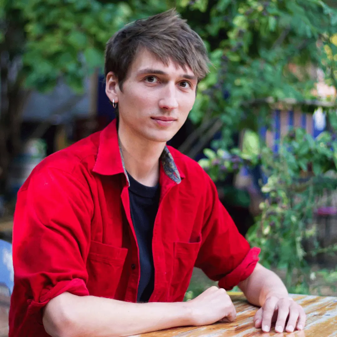 Sur cette image se trouve Charles Deffrennes. Il est assis a une table sur laquelle il s'appuie. Il est vêtu d'une chemise rouge et d'un tee-shirt noir, a les cheveux courts bruns et arbore un sourire. En arrière plan se trouve de la végétation. 