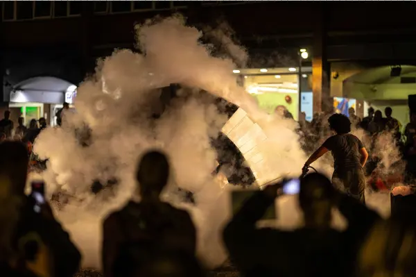 Outside at night. In the foreground audience are silhouetted, some holding up phones, with lit screens, to photograph the performance. A performer fires a fire extinguisher at a completed arch of concrete and ice, creating clouds of vapour which catches the light. Beyond, audiences watch on, silhouetted against the lights of the city behind them. 